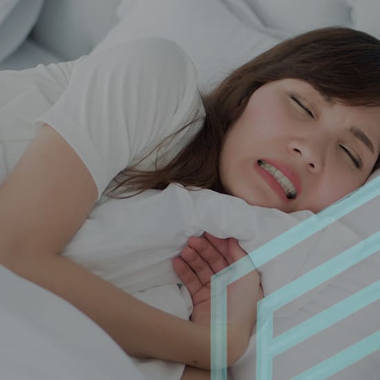 A woman lies in bed with a pained expression on her face, clutching a pillow tightly. She seems to be experiencing discomfort or distress. The background is a white bed with pillows.