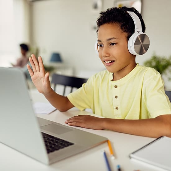 A young boy wearing a yellow shirt and white headphones is smiling and waving while sitting at a desk and looking at a laptop screen. There are notebooks and pencils on the desk, and a blurred background shows another person working.