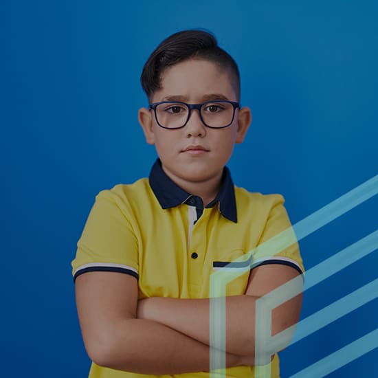 A young boy with short dark hair and glasses stands with a serious expression against a solid blue background. He is wearing a yellow polo shirt with black trim and has his arms crossed.