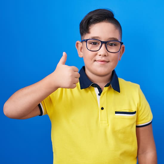 A young boy with short dark hair, wearing glasses and a yellow polo shirt with a black and white collar, gives a thumbs up. He stands in front of a solid blue background.