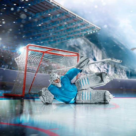 A hockey goalie in blue gear is on the ice in front of the goal net, making a dramatic save with an outstretched arm and stick. The indoor arena is filled with dynamic lighting and a blurred audience in the background. Snow and ice particles enhance the action.