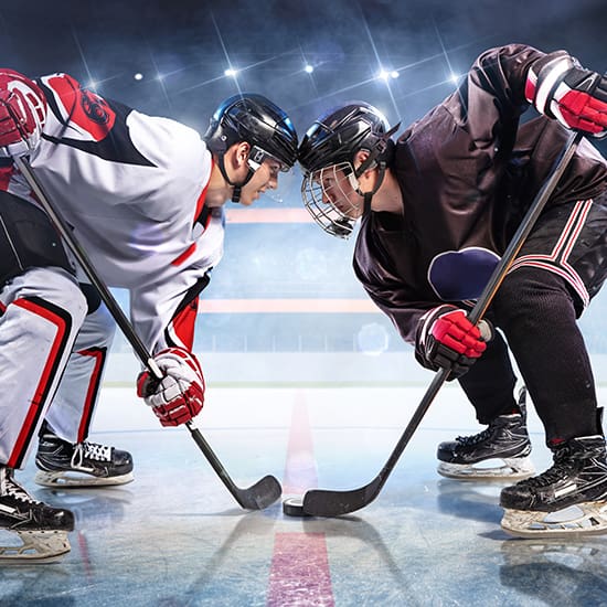 Two ice hockey players face each other on a rink, preparing for a face-off. One player wears a white jersey with red and black accents, and the other wears a black jersey. Both players are bent over with their hockey sticks poised over the puck.