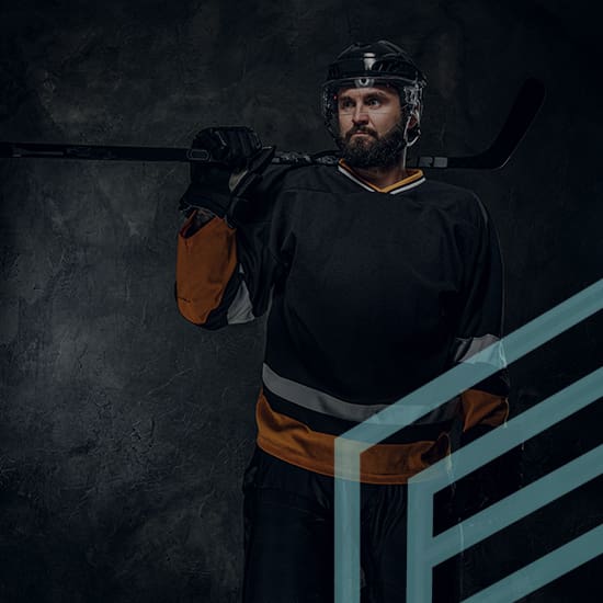 Hockey player standing against a dark background, holding a hockey stick over his shoulder. He is wearing a black jersey with orange accents, protective gear, and a helmet. His expression is serious and focused.