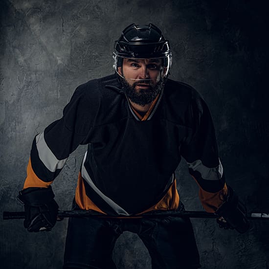 A hockey player wearing a black and orange uniform and a helmet, holding a hockey stick, poses against a dark, textured background. The player has a focused expression and a prominent beard.