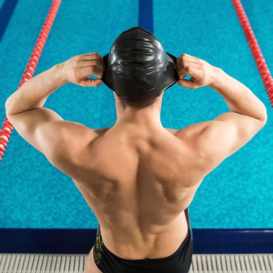 A swimmer stands by the edge of a pool, seen from behind. They are adjusting their black swim cap and are wearing black swim trunks. The person has a muscular build and the pool water is blue, separated by red lane dividers.