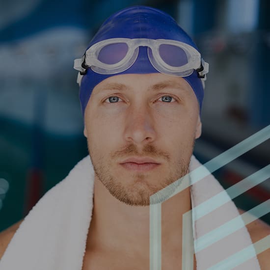 A swimmer wearing a blue swim cap and goggles rests with a white towel around his shoulders. He is looking directly at the camera with a focused expression. The blurred background suggests an indoor pool environment.