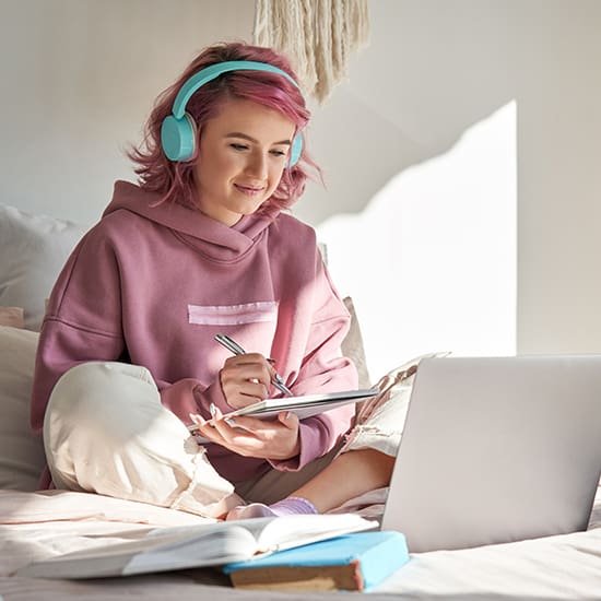 A person with pink hair is sitting on a bed, wearing teal headphones and a pink hoodie. They are taking notes in a notebook while looking at an open laptop. Sunlight streams through a window, and a couple of books lie nearby.