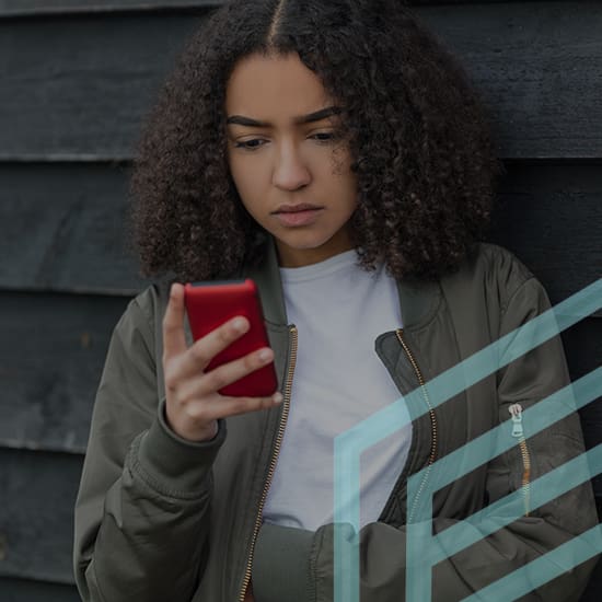A person with curly hair is leaning against a dark wooden wall, holding and looking at a red smartphone with a serious expression. They are wearing a green jacket over a white shirt. A geometric pattern overlay is partially visible on the image.