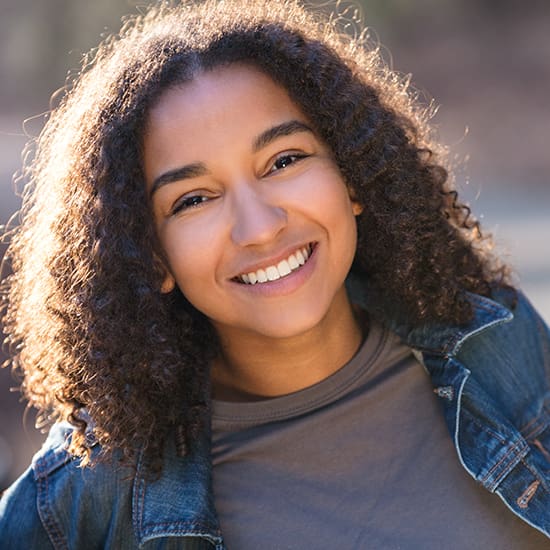 A young person with curly hair smiles warmly at the camera. They are wearing a denim jacket over a t-shirt. The background is softly blurred, focusing attention on their bright expression.