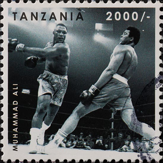 A Tanzania postage stamp features a black-and-white photograph of a boxing match between two fighters in a ring. One boxer, wearing light-colored shorts, appears to be throwing a punch, while the other, in darker shorts, is dodging. Text reads "Tanzania 2000/-" and "Muhammad Ali".