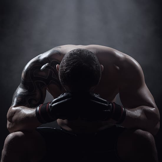 A shirtless man with a tattoo on his arm sits on a stool in a dark, smoky environment. He wears boxing gloves and is hunched over with his elbows resting on his knees, head down, and clasped hands positioned between his knees. The atmosphere conveys intensity.