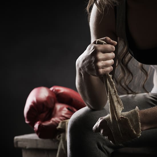 A person is shown wrapping their hands with cloth bandages, preparing for a boxing session. In the background, a pair of red boxing gloves is resting on a wooden bench. The scene is dimly lit and focused on the person's hands and the gloves.