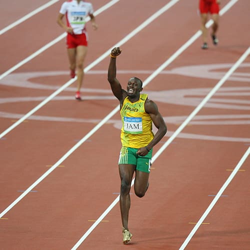 A sprinter wearing a yellow and green uniform, labeled "JAM," raises his fist in celebration as he crosses the finish line on a track. Another athlete is visible in the background. The race appears to be in a large stadium.