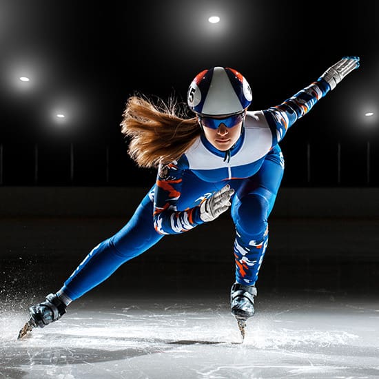 A speed skater dressed in a blue and white skintight suit, helmet, and protective gloves is skating on an ice rink with focused determination. The background is dimly lit with bright overhead lights highlighting the skater's dynamic pose and motion.
