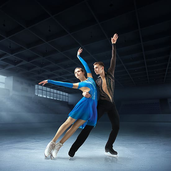 Two figure skaters perform on an ice rink under dim lighting. The female skater wears a blue dress, and the male skater wears black attire. They exhibit graceful postures with arms extended. The empty stadium seats are visible in the background.