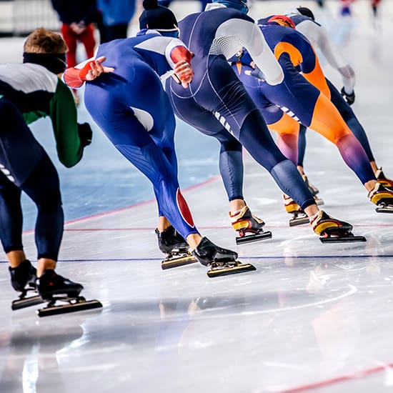 Speed skaters in colorful suits are racing on an indoor ice rink. They are leaning forward in unison as they glide on their skates, creating a dynamic and synchronized movement. The focus is on their lower bodies and skates.
