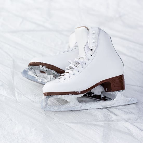 A pair of white figure skates with brown heels rests on an ice surface. The skates have lace-up fronts and are slightly embedded in the ice, with some snow covering the blades. The ice shows faint markings from previous skating activity.