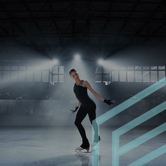 A figure skater performing on an ice rink under bright spotlights. The skater is posing gracefully, wearing a black outfit and gloves, with a spotlight illuminating her from behind. The arena background is dimly lit, with visible stadium seats.