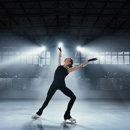 A figure skater in a black outfit strikes a dramatic pose on an ice rink, arms extended upwards. Bright spotlights illuminate the skater, creating an ethereal glow. The background is a dimly lit arena with a grid pattern visible on the ceiling.