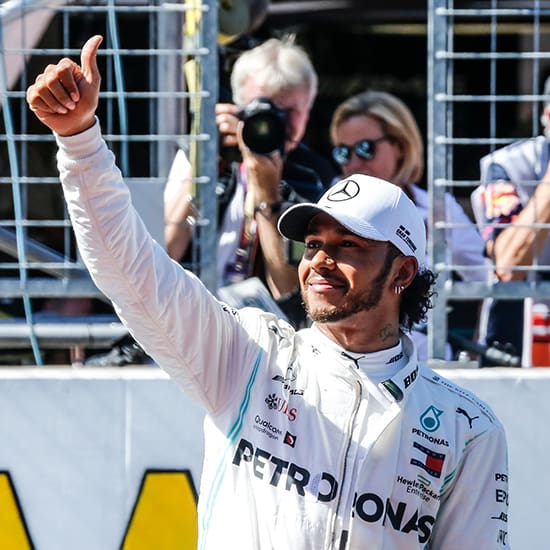A man in a white racing suit and cap gives a thumbs-up while standing on a racetrack. He has a smile on his face, and there are spectators and photographers in the background. The suit has various sponsor logos, including Petronas.