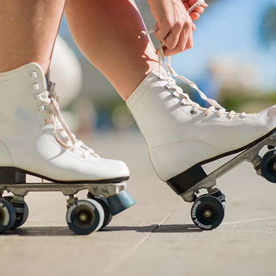 Close-up of a person wearing white roller skates, adjusting the laces. The background is slightly blurred, showing an outdoor scene with bright sunlight. The person is kneeling, and only their hands and legs are visible.