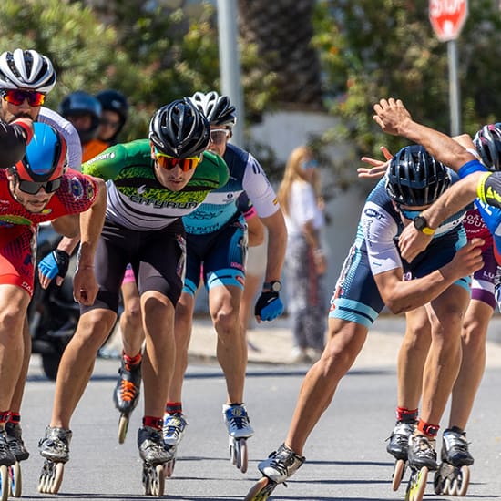 A group of inline skaters in colorful racing gear competes in an outdoor event on a sunny day. They are leaning forward and closely packed as they navigate a turn. In the background, spectators watch near a stop sign and palm trees are visible.