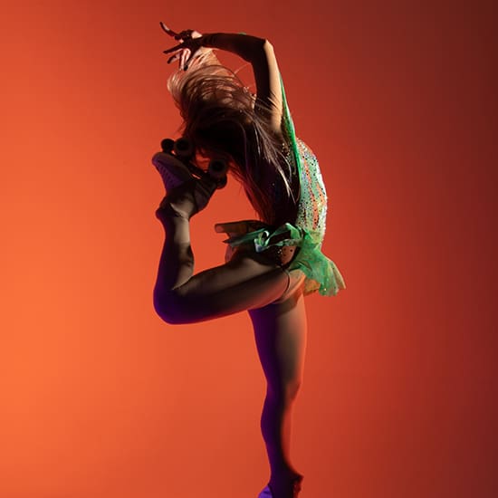 A roller skater in a green sequin outfit performs a high-energy jump against a vibrant orange background. The skater's arms are gracefully extended, and one leg is bent, showing dynamic movement and athleticism.