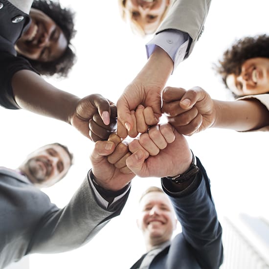 A diverse group of people facing inward, gathering their hands in a circle and doing a fist bump. They are smiling, wearing business attire, and the picture is taken from a low angle against a bright sky.