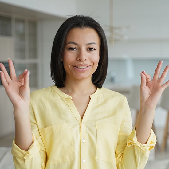 A woman with shoulder-length dark hair, dressed in a yellow shirt, is smiling and holding her hands up, making an "OK" gesture with both hands. She is standing in a bright, modern kitchen.