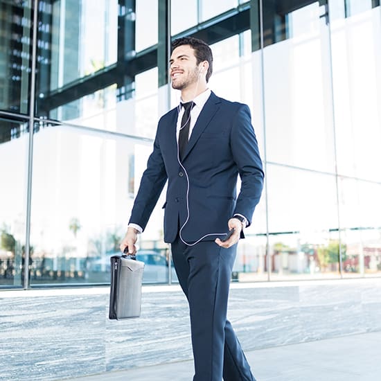 A man dressed in a navy blue suit, white shirt, and tie walks outside a modern glass building. He is smiling, holding a briefcase in his left hand, and has earphones in his ears. The background reflects the cityscape and sky.