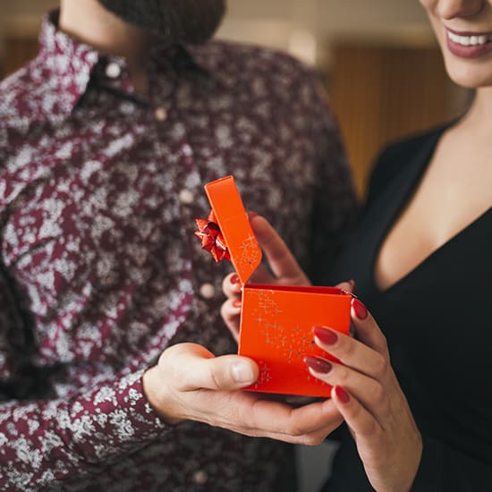 A close-up of a couple holding a small, opened red gift box adorned with a red ribbon. The person's fingernails are painted red, matching the gift box. The couple appears to be smiling. One person is wearing a patterned shirt, and the other is in a black top.
