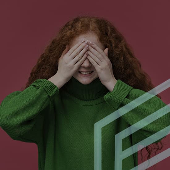 A person with long, curly red hair is smiling and covering their eyes with their hands. They are wearing a green sweater and standing in front of a burgundy background. The image features geometric patterns in the bottom right corner.