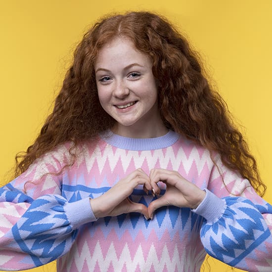 A young person with long, curly red hair smiles at the camera while making a heart shape with their hands. They are wearing a sweater with a geometric pattern in pink, blue, and white. The background is bright yellow.