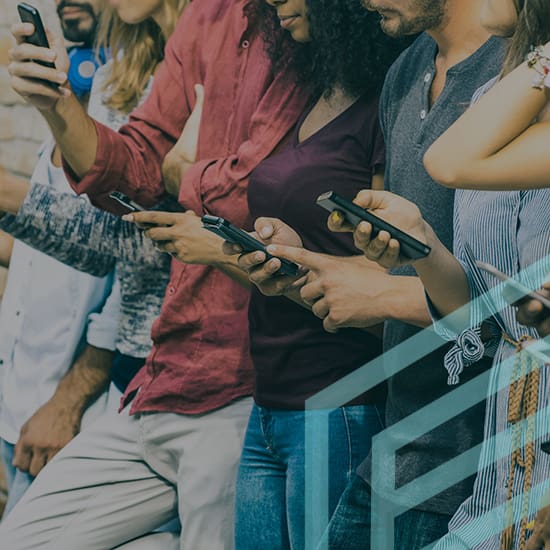 A group of people standing closely together, all looking down at their smartphones. They are dressed casually, and their focus is entirely on their devices. Only parts of their bodies and devices are visible, not their full faces.