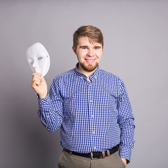 A man with short hair and a beard is wearing a blue and white checkered shirt and khaki pants. He is standing against a gray background, smiling, and holding a white mask in his right hand.