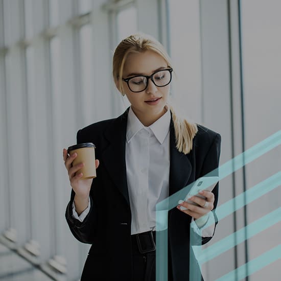 A woman with blonde hair wearing glasses, a white shirt, and a black blazer is holding a coffee cup in one hand and looking at her smartphone in the other. She is standing in a modern, well-lit space with large windows.