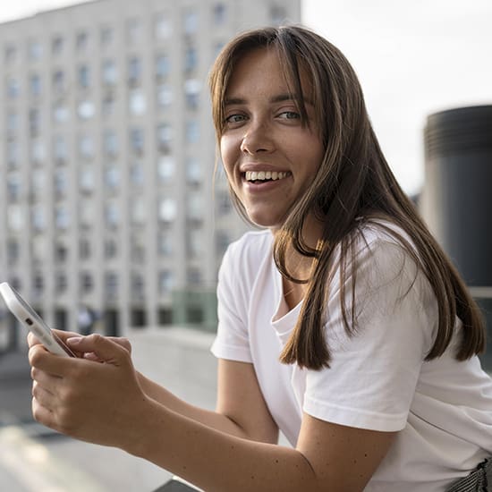 A young woman with long brown hair and a white shirt is holding a smartphone and smiling at the camera. She is standing outdoors in an urban area with a large building in the background.