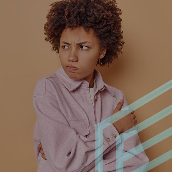 A young person with curly hair frowns and looks to the side while crossing their arms. They are wearing a light pink jacket and standing against a solid beige background with a faint blue geometric pattern overlaying part of the image.