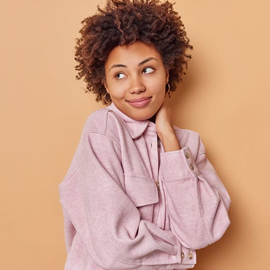 A person with curly hair smiles and looks off to the side. They are wearing a light pink, long-sleeved shirt and have their hand resting on their neck. The background is a plain, light brown color.