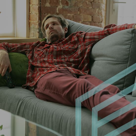 A person in a red plaid shirt and red pants is lounging on a gray sofa with arms outstretched over the backrest. They are holding a remote control and appear to be relaxing in a room with a brick wall and windows.