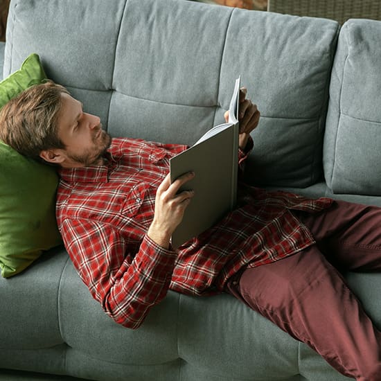 A man with a beard, wearing a red plaid shirt and maroon pants, is lying on a gray sofa reading a book. A green pillow supports his head as he relaxes comfortably on the couch. The background shows part of a wicker chair.
