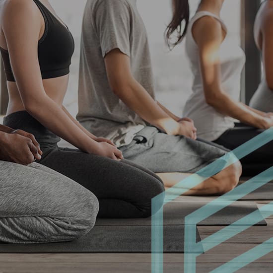 Four people are sitting cross-legged on yoga mats indoors, with minimalistic and bright decor in the background. They are wearing comfortable athletic clothing, and their hands are resting on their knees in a relaxed position. The focus is from the shoulders down.