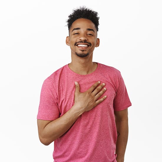 A smiling person with short curly hair is standing against a plain white background, wearing a pink t-shirt. They have one hand placed over their chest, conveying a friendly and appreciative gesture.