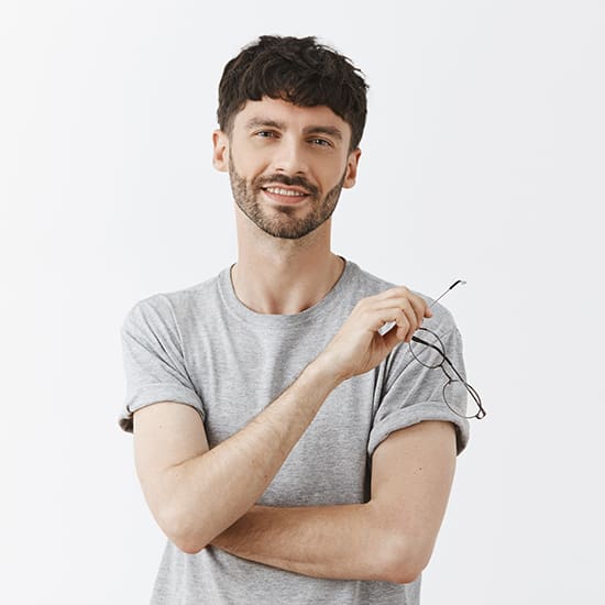 A smiling man with short, wavy dark hair and a beard is standing against a plain white background. He is holding a pair of eyeglasses in his right hand and has his left arm crossed over his chest. He is dressed in a light grey rolled-up sleeved T-shirt.