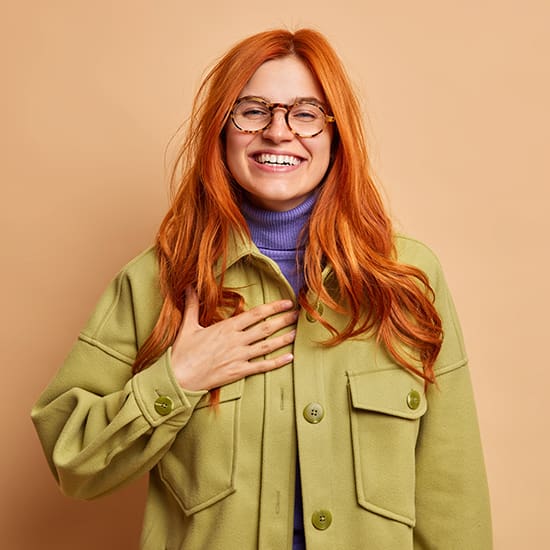 A person with long red hair, wearing glasses, a green jacket, and a purple turtleneck, smiles warmly with one hand placed over their chest. The background is a soft peach color.