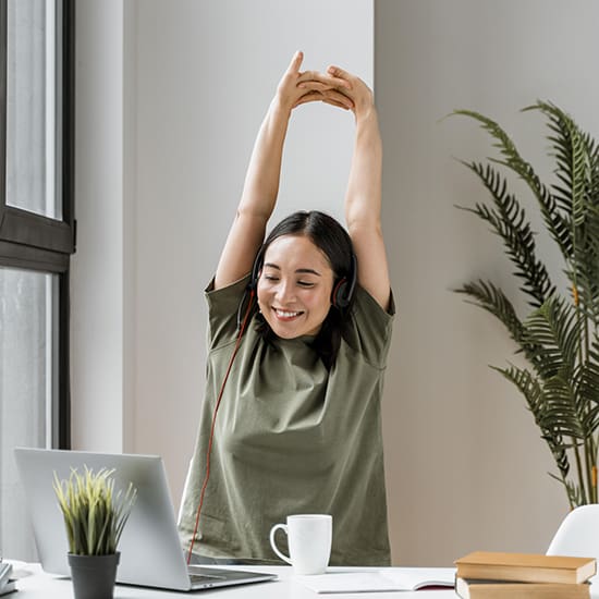 A woman wearing headphones and a green shirt is sitting at a desk with a laptop, plant, and coffee mug, stretching her arms above her head. She is smiling and appears relaxed. A large plant and a window are in the background.