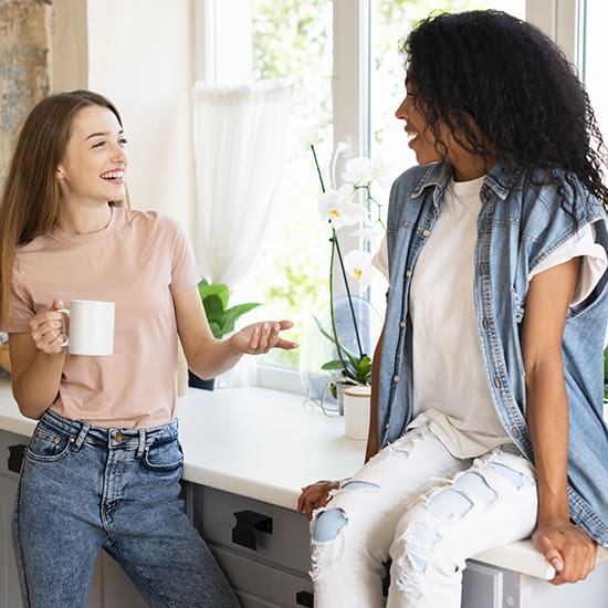Two women are conversing and smiling while one sits on a counter and the other stands holding a mug. The woman standing wears a light pink shirt and jeans, the seated woman wears a white shirt and denim jacket. They are in a well-lit room with a window and plants.