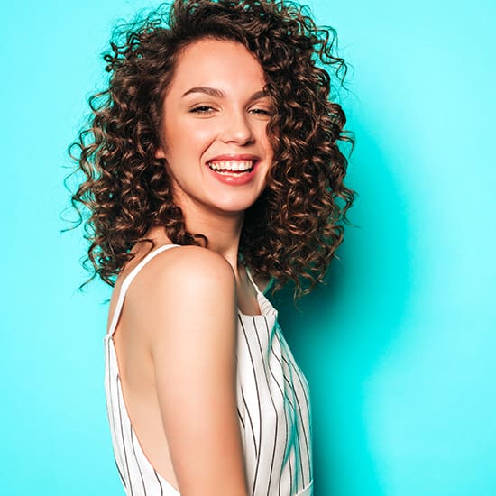 A young woman with curly hair stands against a vibrant turquoise background. She is wearing a sleeveless, white dress with vertical black stripes. She is smiling and looking towards the camera, exuding a joyful and confident demeanor.
