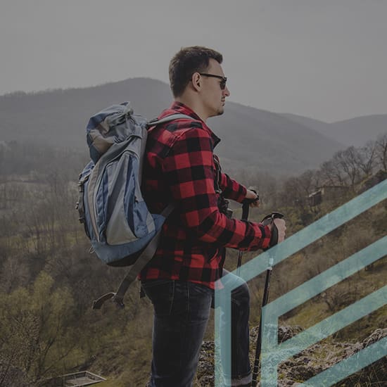 A man wearing a red and black plaid shirt, carrying a backpack, and holding trekking poles stands on a scenic hillside. The landscape features lush greenery and distant, misty mountains. He faces sideways, gazing into the distance.