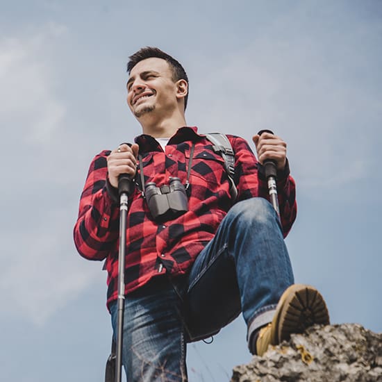 A man wearing a red and black checkered shirt, jeans, and hiking boots stands on a rocky surface. He's holding trekking poles and has binoculars around his neck. He looks content, gazing into the distance with a clear sky in the background.
